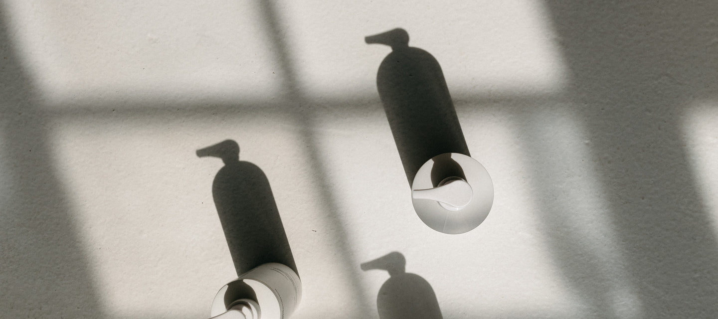 Four white bottles with pointed caps are arranged on a textured surface, casting long shadows. Sunlight streams through a window, creating a grid pattern of light and shadow across the scene.