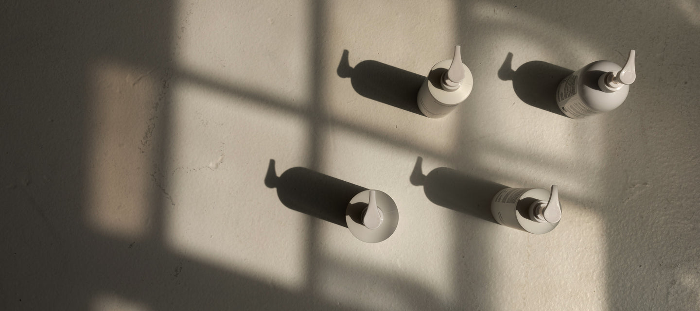 Four minimalist bottles with droppers cast long shadows on a textured, light-colored surface, illuminated by soft sunlight filtering through a window grid.