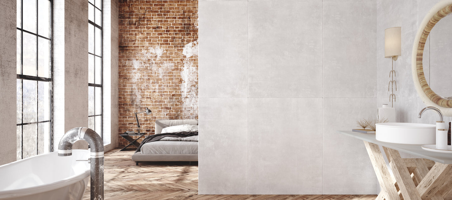 A modern loft with a freestanding bathtub, wooden herringbone floor, large windows, exposed brick walls, and a cozy bed partially visible in the adjoining room.