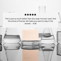 A beige bar of The Cleansing Clay by Basic Maintenance rests on a reflective surface amid glassware filled with clear liquid. Above, a five-star review highlights the soap’s exceptional quality and aroma.