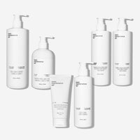 Six white pump bottles and tubes from Basic Maintenances Total Transformation Kit are arranged against a white background, each labeled with different skincare product types in minimalist black text.