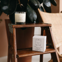 The Calm candle by Basic Maintenance, in its white box, sits on a wooden step ladder with a woven chair and large leafy plant in the background.