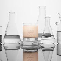 The Calm candle by Basic Maintenance sits in a glass jar among clear laboratory beakers and flasks with liquids, arranged on a reflective surface against a white background.