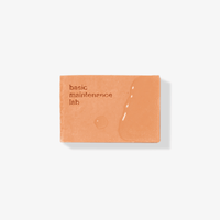 A rectangular orange bar of The Cleansing Clay by Basic Maintenance rests on a white background, featuring the brand name engraved and a few water droplets on its surface.