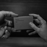 Two hands exchange a small rectangular box labeled The Cleansing Clay from Basic Maintenance; the image is in black and white.