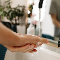 A close-up shows hands lathered with The Hand Soap by Basic Maintenance under a black faucet, running water rinsing away suds. In the blurred background, a plant and bathroom essentials sit near the sink.