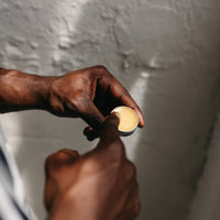 A person with dark skin scoops The Lip Fix balm by Basic Maintenance from a small round metal tin against a light, textured background.