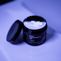 A black jar of The Protein Pomade by Basic Maintenance sits open on a smooth surface under bluish-purple light, revealing white cream inside.